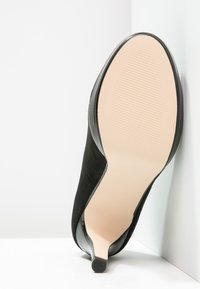 Černá semišová vysoká podpatková bota s lesklým černým obrysem a texturovanou béžovou podrážkou, nakloněná proti bílému pozadí.