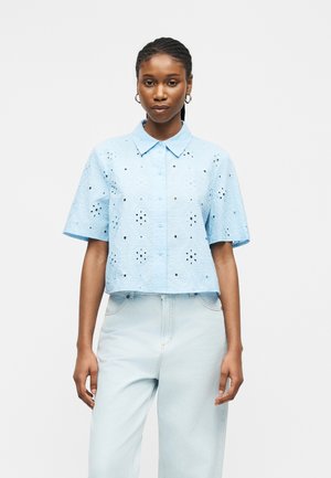 Žena odjevena u svijetloplavu kratku košulju s rupicama i svijetloplave traperice s visokim strukom na jednobojnoj pozadini.