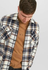 Camisa de franela a cuadros en azul, beige y blanco con dos bolsillos en el pecho, cierre de botones frontal y una camiseta de cuello redondo beige debajo.