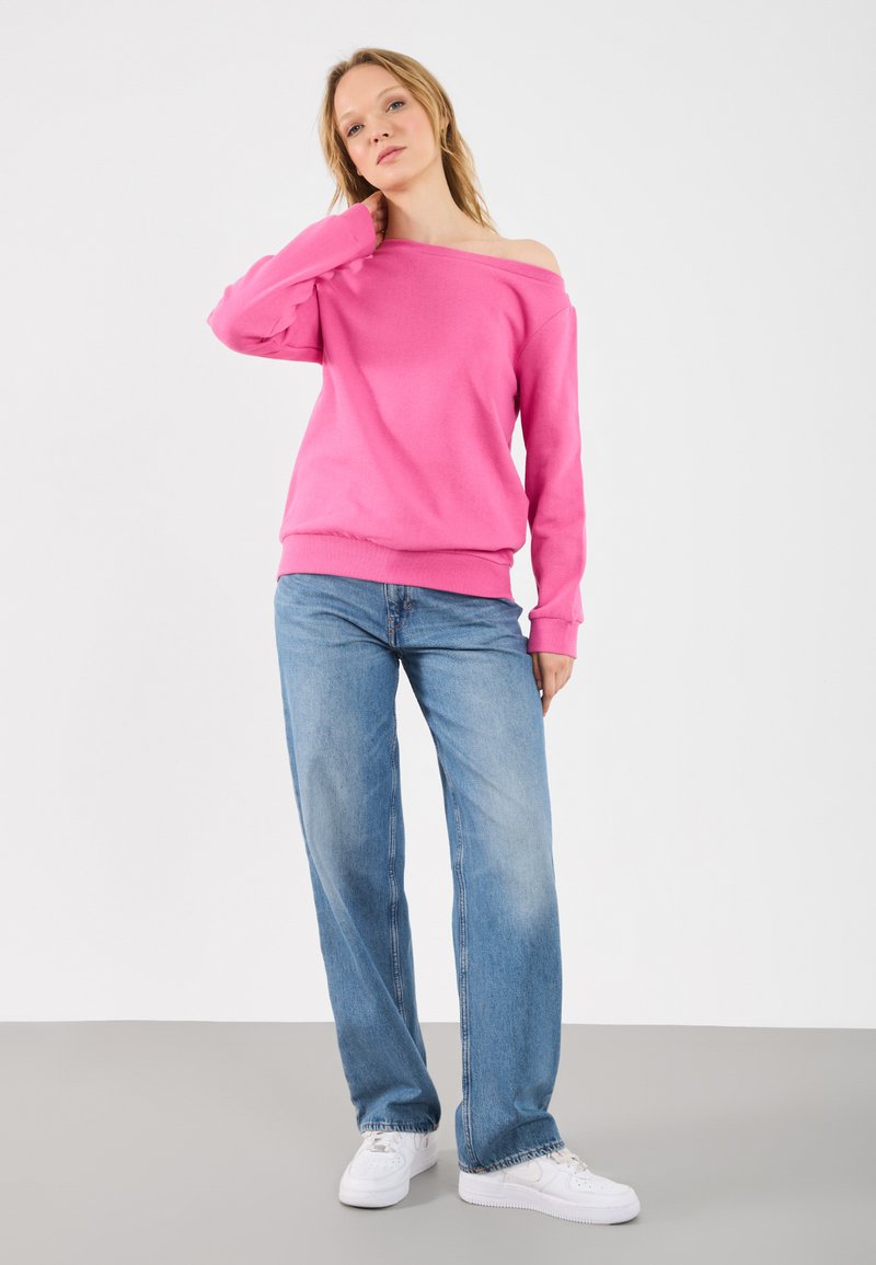 Różowy sweter typu off-shoulder wykonany z miękkiego materiału; w parze z jasnoniebieskimi spodniami o szerokich nogawkach i białymi sneakersami. Minimalistyczne tło.