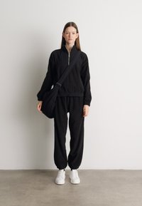 Femeie îmbrăcată într-o bluză neagră cu fermoar și pantaloni, încălțăminte albă, purtând o geantă neagră tip crossbody, stând în fața unui perete alb și a unei podele gri.