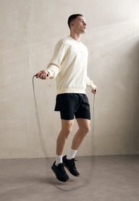 Lys beige sweatshirt, sorte atletiske shorts og sorte sneakers. Personen hopper i sjippetov, med en neutral baggrund og blødt lys.