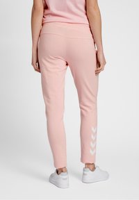 Person som bär ljusrosa joggers med vita chevron-ränder vid ankeln och vita sneakers, stående mot en enfärgad grå bakgrund.