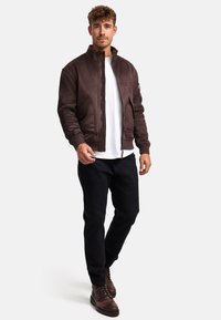Chaqueta bomber de ante marrón con un cuello alto, cremallera frontal y bolsillos laterales, combinada con una camiseta blanca y pantalones oscuros. Botas de cordones marrones.