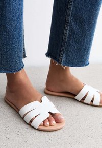 Sandalias planas blancas con un diseño de tiras anchas, textura suave y suela tostada, combinadas con unos vaqueros azules de dobladillo deshilachado sobre una superficie clara.