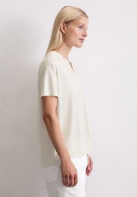T-shirt henley crème à manches courtes avec une coupe décontractée, présentant un col rond et cinq boutons à l'avant. Texture lisse.