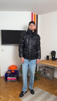 Čierna nadýchaná bunda so lesklou textúrou, kapucňou a prednými vreckami, nosená nad svetlo modrými jeansami a čiernou obuvou. Minimálne detaily z kovu.