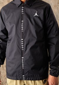 Černá větrovka s límcem, vybavená středovým zipem a bílým nápisem "Jordan" na přední straně, vedle loga Jumpman na hrudi.