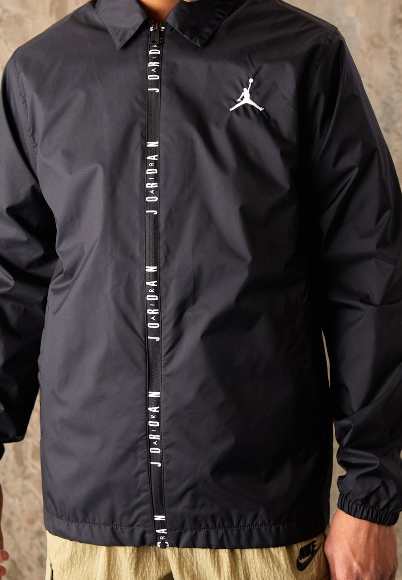 Černá větrovka s límcem, vybavená středovým zipem a bílým nápisem "Jordan" na přední straně, vedle loga Jumpman na hrudi.