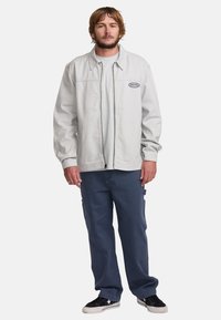 Light gray denim jacket with a zip front, side pockets, and a black logo patch. Paired with dark blue cargo pants and black sneakers.
