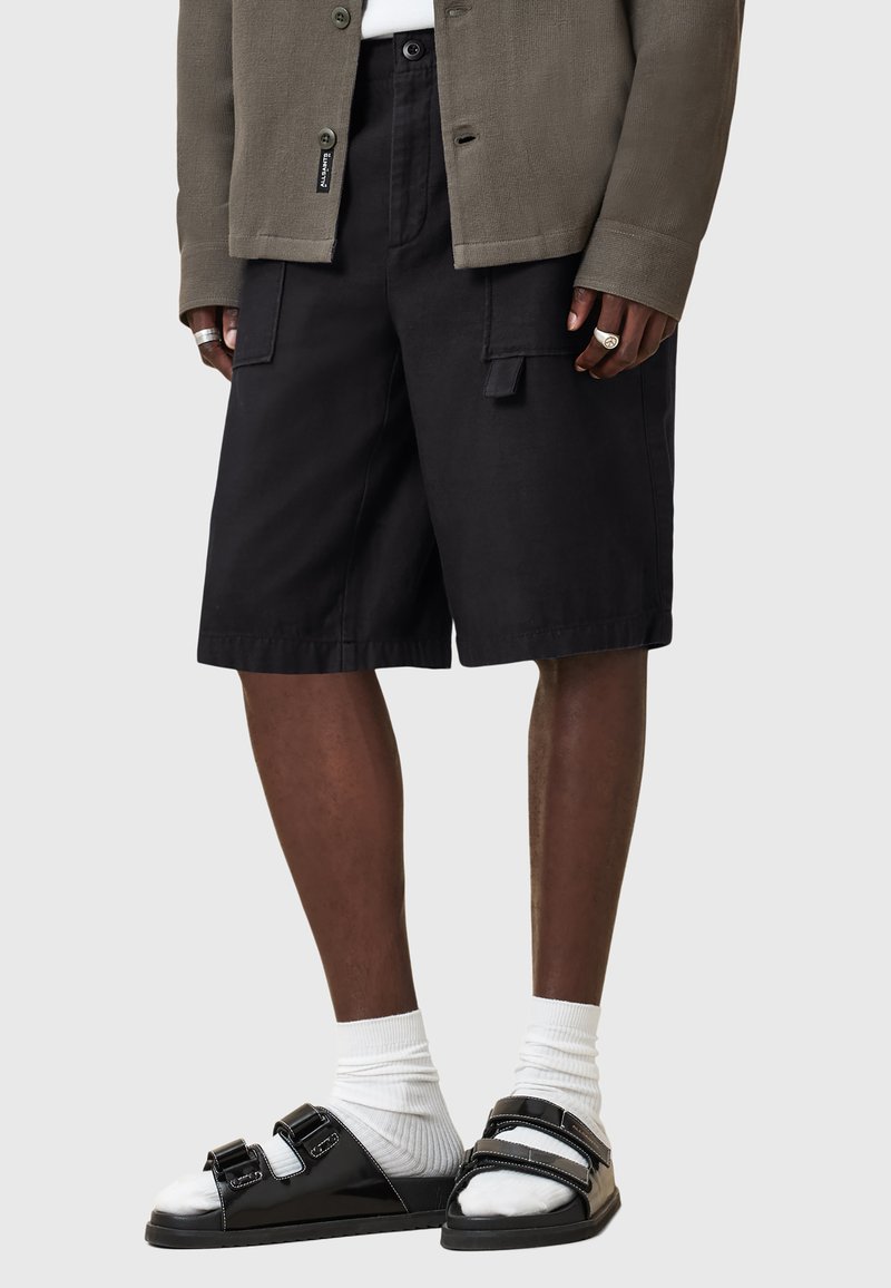 Black cargo shorts with front pockets, paired with black sandals featuring a shiny finish and adjustable straps over white socks. Textured grey jacket.
