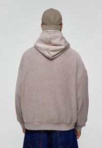 Lichtgrijze oversized hoodie met een zachte textuur, voorzien van een trekkoord capuchon en geribde manchetten. Gecombineerd met een beige pet.