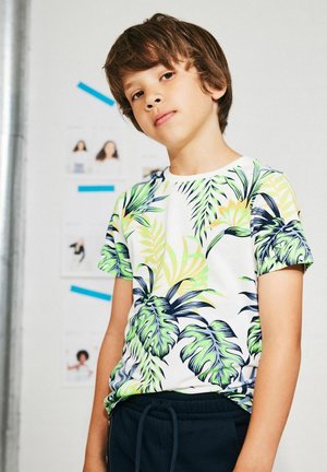 Camiseta de manga corta con fondo blanco y un vibrante estampado de hojas tropicales en verde y amarillo, combinada con pantalones oscuros con cordón.