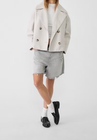 Double-breasted, light gray wool jacket with large brown buttons, paired with frayed gray denim shorts and classic black shoes with white socks.