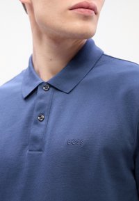 Hombre vestido con una camiseta polo azul con cuello y dos botones, que presenta un pequeño logo bordado de "BOSS" en el pecho.