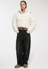Przycięta, puszysta beżowa kurtka z kapturem i elastycznymi mankietami, zestawiona z szerokimi, ciemnymi dżinsami. Brązowe obuwie dopełnia stylizację.