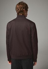 Jachetă bomber maro închis cu guler ridicat, manșete elastice și tiv. Fabricată dintr-un material neted, prezintă un design curat și minimalist.