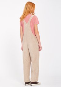 Beige kordfløjlsoveralls med brede ben, justerbare stropper, sidelommer og en afslappet pasform, parret med en pink T-shirt og brune sneakers.