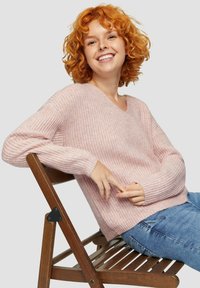 Maglione rosa chiaro a maglia con scollo a V e maniche a costine. Il modello è seduto su una sedia di legno, indossando jeans in denim. Texture morbida visibile.