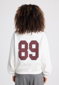 White sweatshirt with a relaxed fit, featuring a large maroon "89" on the back, outlined in light blue. Soft, textured fabric.