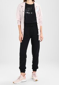 Sportovní outfit značky Nike zahrnující světle růžovou větrovku, černé tričko s logem, černé tepláky a růžové tenisky s bílými detaily.