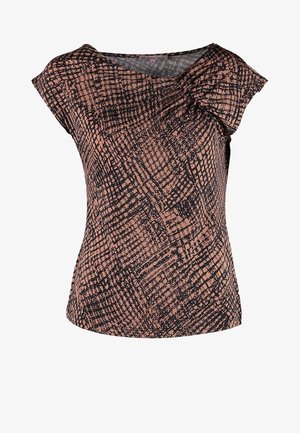 La femme porte un haut à manches courtes marron et noir à motifs, un jean skinny foncé, des bottines noires, et tient un sac fourre-tout noir dans sa main gauche.