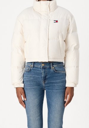 Frau trägt eine weiße, kurze Steppjacke mit hohem Kragen und blaue Jeans, steht mit entspannt an den Seiten hängenden Armen.