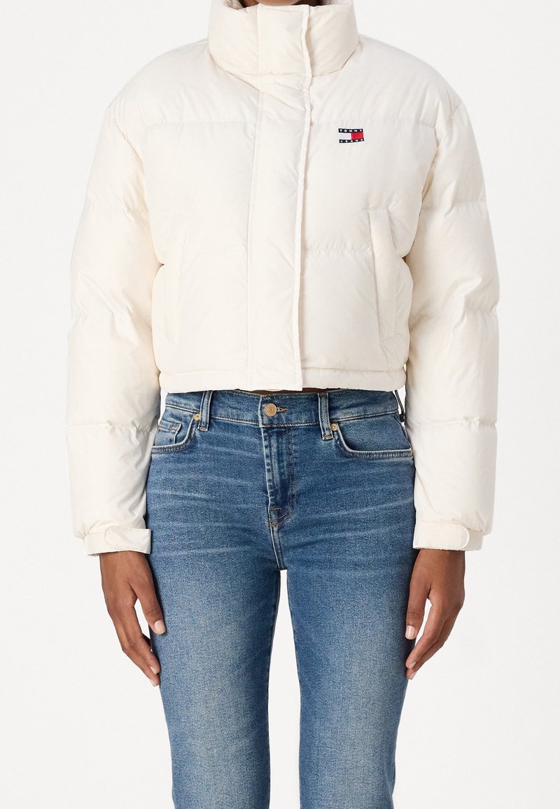 Femme portant une doudoune courte blanche à col montant et un jean bleu, debout avec les bras détendus le long du corps.