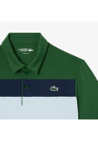 Grön och blå randig polo tröja gjord av andningsbart tyg, med krage, tre knappar och en Lacoste-logga på bröstet.