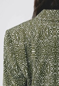 Olivgrön kavaj med intrikata vita geometriska mönster. Texturerat tyg, vikkant och synliga axelsömmar.