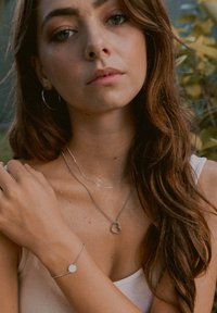 Silberne Schmuckstücke mit einem herzförmigen Anhänger und einem runden Charme an einer Kette, getragen mit einem ärmellosen Oberteil und strukturiertem Haar.