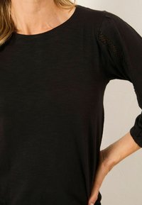 Nahaufnahme einer Person, die ein schwarzes Langarmshirt mit Spitzenverzierungen an den oberen Ärmeln trägt, mit einer Hand auf der Hüfte.