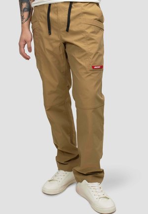 Beige Cargohose mit mehreren Taschen und einem roten "JACKIT"-Etikett am rechten Oberschenkel, getragen mit weißen Sneakern und einem sichtbaren tätowierten Arm.