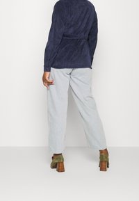 Marinblå mockajacka med midjeband, kombinerad med ljusgrå vida byxor och gröna klackskor, visad från baksidan.