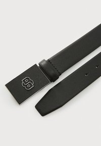 Zwarte synthetische riem met een gestructureerd oppervlak, voorzien van een rechthoekige zilveren logo-buckle en vijf verstelbare gaten voor maatvoering.