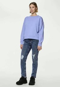 Ανοιχτό μωβ φούτερ με στρογγυλή λαιμόκοψη και ριμπ μανσέτες, σε συνδυασμό με μπλε παντελόνι τζιν με φθαρμένα σημεία και μαύρα αθλητικά παπούτσια.