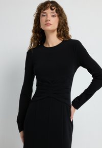 Schwarzes langärmeliges Kleid mit tailliertem Oberteil, gerafften Details an der Taille und geradem Rock. Glatter Stoff und minimalistisches Design.