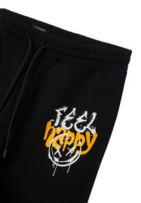Pantaloni della tuta neri con vita elasticizzata e coulisse. Presentano la scritta "feel happy" in stampa stile graffiti bianca e arancione, e una grafica di un volto sorridente.