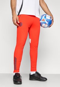 Pantalones deportivos rojos con diseño ajustado, que presentan un logo del Bayern de Múnich, bolsillo lateral y rayas azul marino a los lados. Zapatos de fútbol negros.