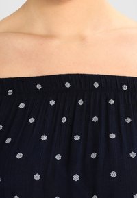Top blu navy scollato con tessuto texturizzato, caratterizzato da ricami geometrici bianchi e un collo arricciato.