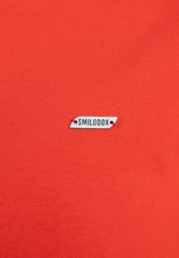 Camiseta de algodón roja con una pequeña etiqueta plateada que lleva el texto "SMILODOX" unida con hilo rojo. Textura suave con un diseño ajustado.
