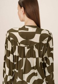 Femme aux longs cheveux bruns portant une chemise ample à manches longues avec des motifs géométriques abstraits verts foncés et blancs, vue de dos.