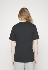 Person med långt ljusbrunt hår som bär en enkel svart t-shirt med kort ärm och ljusgrå byxor, stående mot en vit bakgrund.