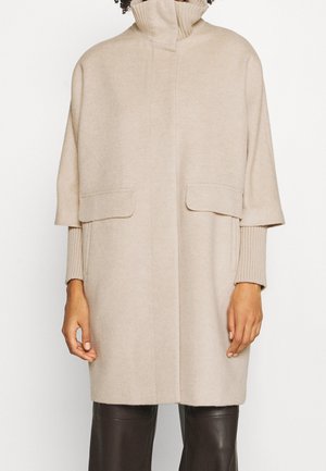 Manteau beige mi-longueur aux genoux avec un col montant, des manches courtes larges sur des manches longues côtelées, et des poches à rabat, porté avec un pantalon foncé.