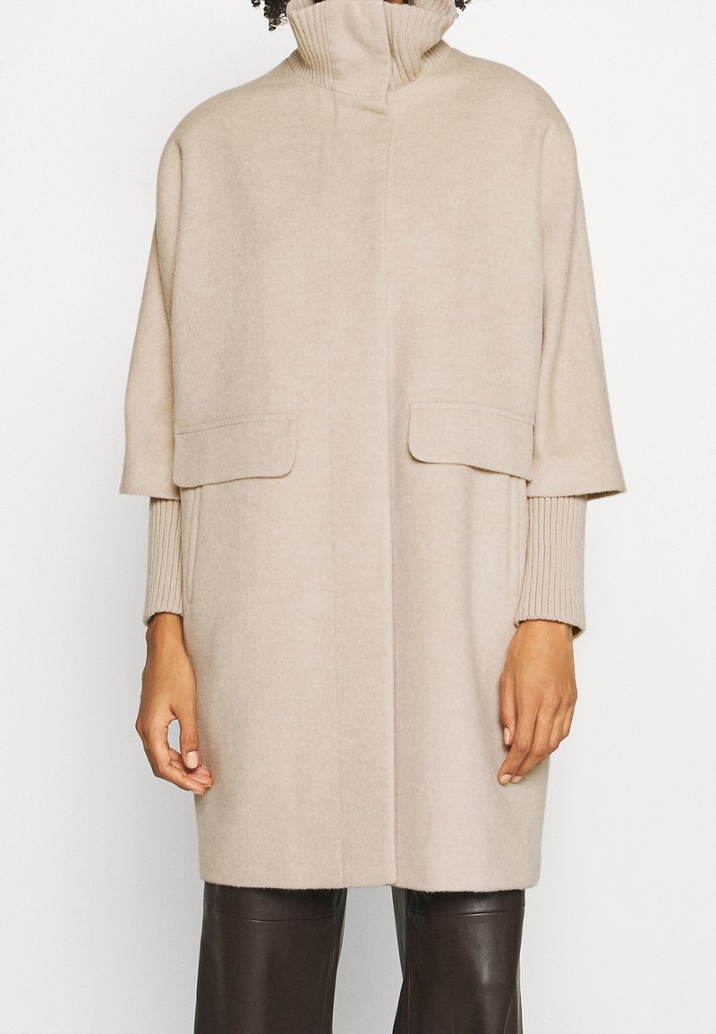 Manteau beige mi-longueur aux genoux avec un col montant, des manches courtes larges sur des manches longues côtelées, et des poches à rabat, porté avec un pantalon foncé.