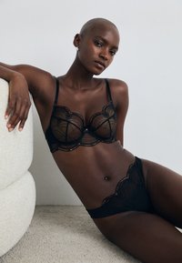 Moteris su nuskustais plaukais, dėvinti juodą nėrinį liemenėlę, viena ranka remiasi į tekstūruotą šviesios spalvos kėdę, sėdinti ant smėlio spalvos kilimo.