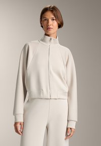 Vaalean beige vetoketjullinen takki, jossa on korkea kaulus, ribattu pinta ja raglan-hihat; yhdistettynä samaan sävyyn ribattuihin housuihin.