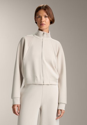 Light beige zip-up jacket with a high collar, ribbed texture, and raglan sleeves; paired with matching ribbed pants.