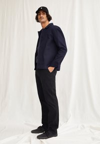 Donker marineblauwe jas met twee voorkruiken, gecombineerd met zwarte broek en donkere schoenen, gedragen door een persoon met krullend haar en een zwarte pet.