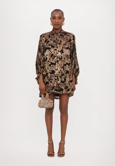 Rochie cu model paisley negru și auriu, cu guler, mâneci lungi și detalii plisate. Modelul ține o geantă mică, cu un model.